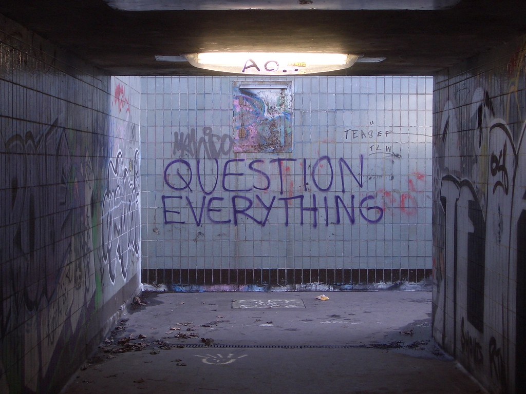 "Question Everything" spray-painted as graffiti in a tiled pedestrian underpass